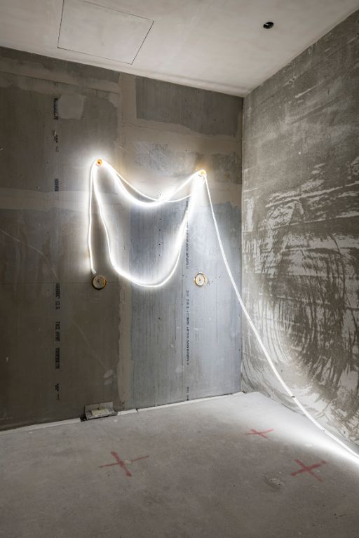 Unfertiger Raum mit rohem Betonboden und kahlen Wänden. Ein leuchtendes Kabel hängt wellenförmig an der Wand und wirft ein sanftes Licht auf den Boden.